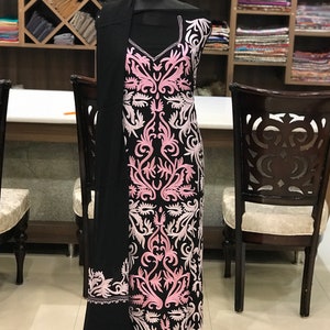 May include: A black sleeveless tunic featuring a V-neckline and intricate light pink and white floral embroidery patterns covering the front. A matching black shawl with a pink embroidered border is draped over the shoulder of the garment.