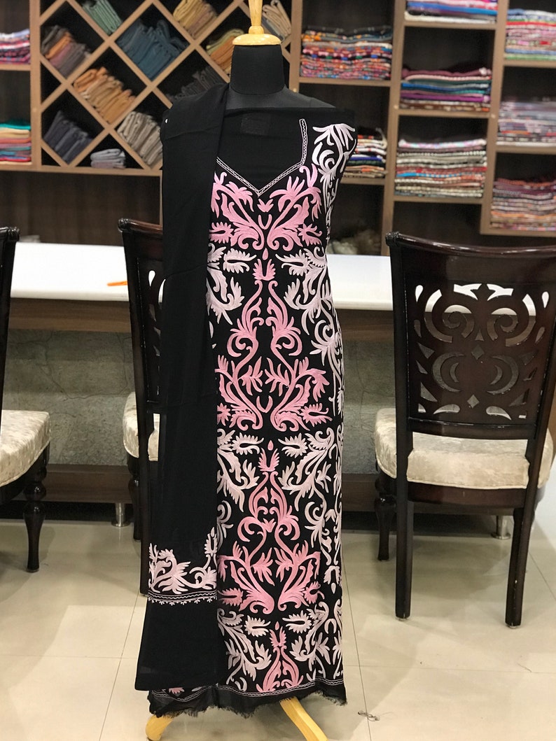 May include: A black sleeveless tunic featuring a V-neckline and intricate light pink and white floral embroidery patterns covering the front. A matching black shawl with a pink embroidered border is draped over the shoulder of the garment.