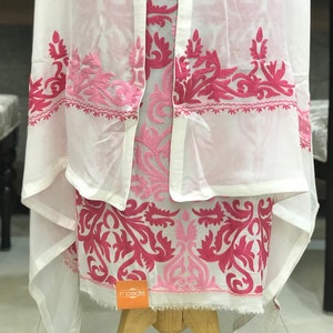 May include: A white garment featuring intricate hot pink and light pink floral embroidery. The fabric is sheer and lightweight, with a draped silhouette and a small orange square tag with the text r'cade visible at the bottom hem.