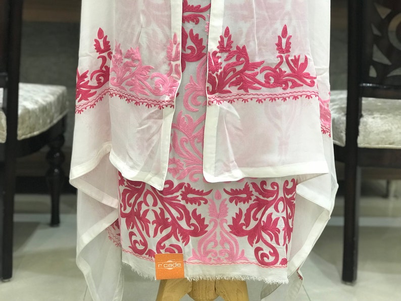 May include: A white garment featuring intricate hot pink and light pink floral embroidery. The fabric is sheer and lightweight, with a draped silhouette and a small orange square tag with the text r'cade visible at the bottom hem.