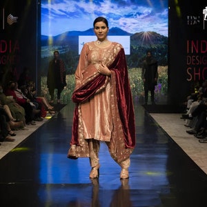 May include: A peach-colored salwar kameez set featuring intricate gold embroidery on the tunic and trousers, paired with a deep maroon velvet shawl draped over the shoulders. The ensemble includes a long-sleeved tunic and matching tapered pants.