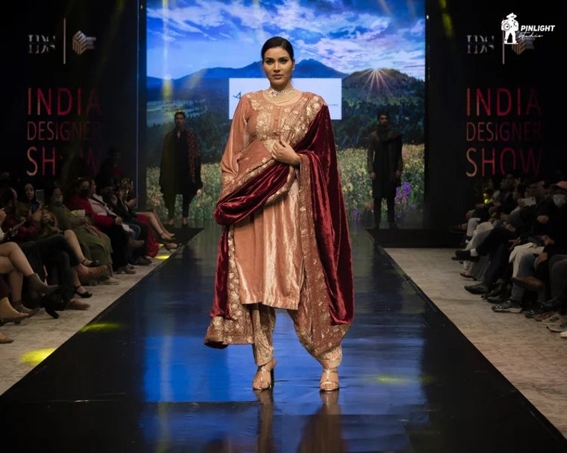 May include: A peach-colored salwar kameez set featuring intricate gold embroidery on the tunic and trousers, paired with a deep maroon velvet shawl draped over the shoulders. The ensemble includes a long-sleeved tunic and matching tapered pants.