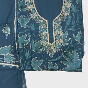 May include: A dark teal fabric garment featuring intricate embroidery of leaves and vines in shades of teal and light gold. The neckline is framed by a decorative gold-toned border, and the pattern extends across the front of the piece.