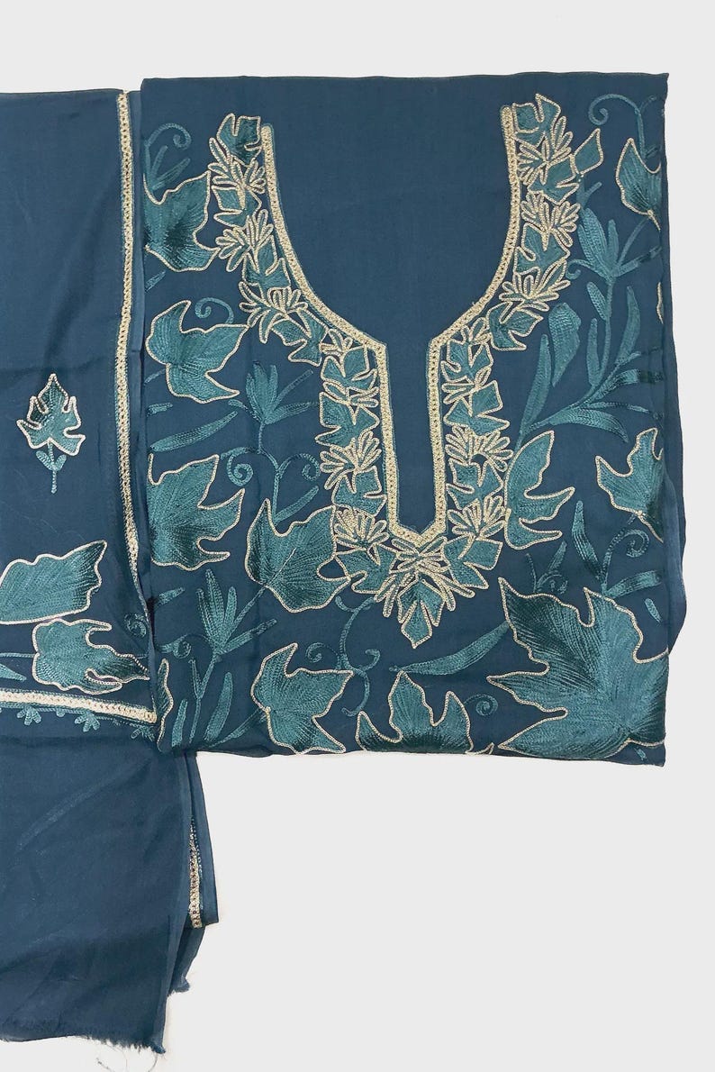 May include: A dark teal fabric garment featuring intricate embroidery of leaves and vines in shades of teal and light gold. The neckline is framed by a decorative gold-toned border, and the pattern extends across the front of the piece.
