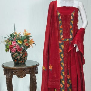 May include: A sleeveless red salwar kameez set featuring intricate embroidery with dark green, orange, and yellow floral and paisley patterns. The ensemble includes a matching red dupatta with delicate embroidered accents along the edges.