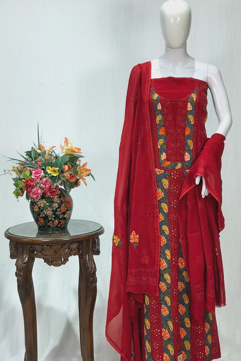 May include: A sleeveless red salwar kameez set featuring intricate embroidery with dark green, orange, and yellow floral and paisley patterns. The ensemble includes a matching red dupatta with delicate embroidered accents along the edges.