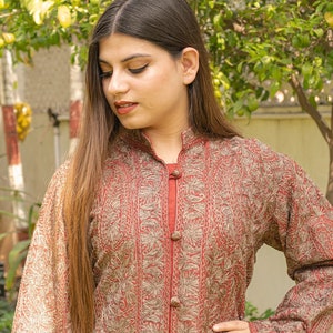May include: A woman wearing a long-sleeved, maroon-coloured tunic with a gold embroidered pattern. The tunic has a mandarin collar and a button-up front.