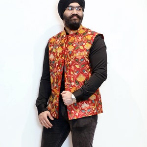 Op de afbeelding: Een man draagt een rood vest met bloemenprint over een zwart shirt en een zwarte jeans. Hij draagt een zwarte tulband en een bril.