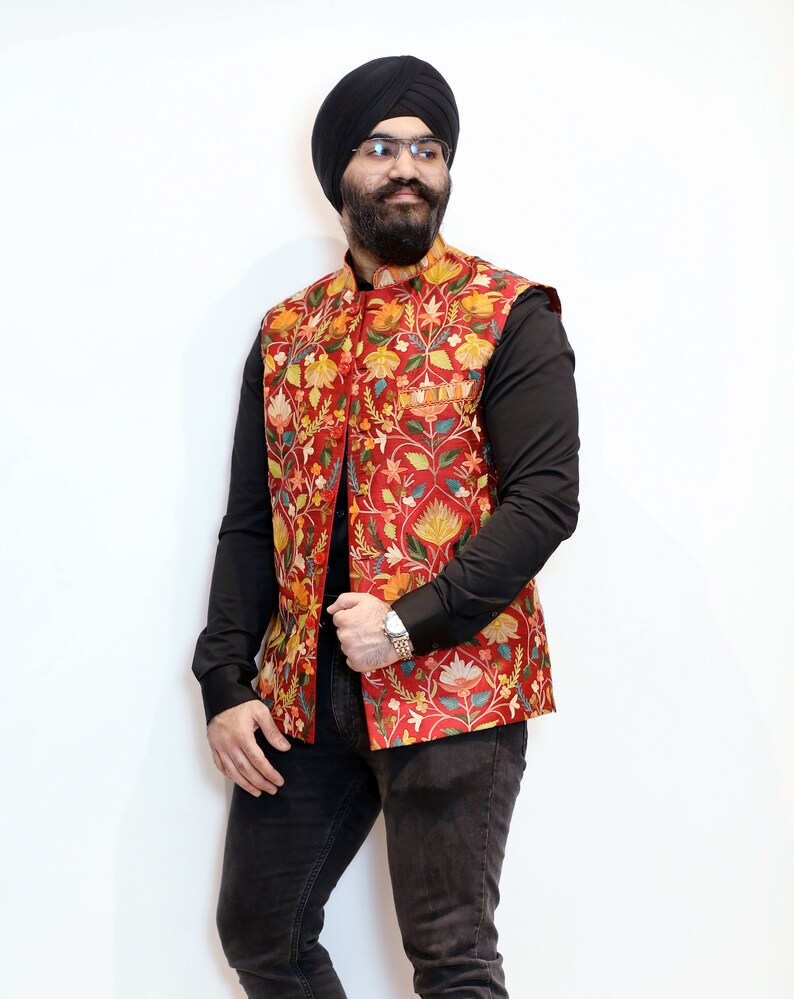 Op de afbeelding: Een man draagt een rood vest met bloemenprint over een zwart shirt en een zwarte jeans. Hij draagt een zwarte tulband en een bril.