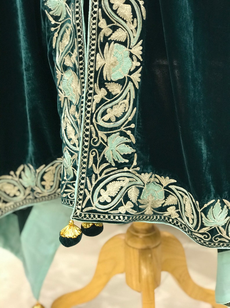 May include: A teal velvet shawl with a delicate floral embroidery pattern in silver and light blue. The shawl has a fringe trim and tassels.