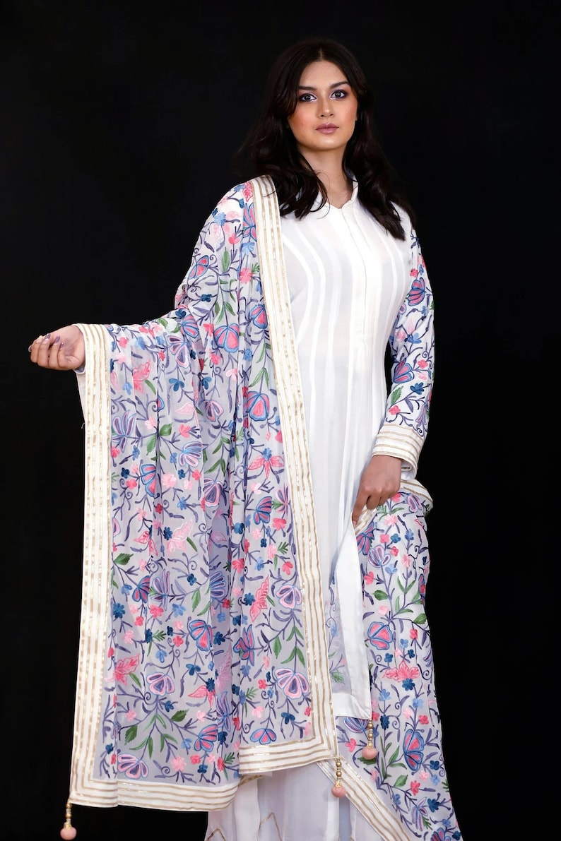 May include: A white embroidered dupatta with a floral pattern in shades of pink, blue, and green. The dupatta has a gold and white border and a tassel at the end.