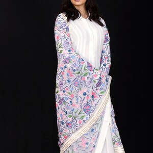 May include: A woman wearing a white long-sleeved shirt with a white dupatta featuring a floral embroidery pattern in shades of pink, blue, and green.