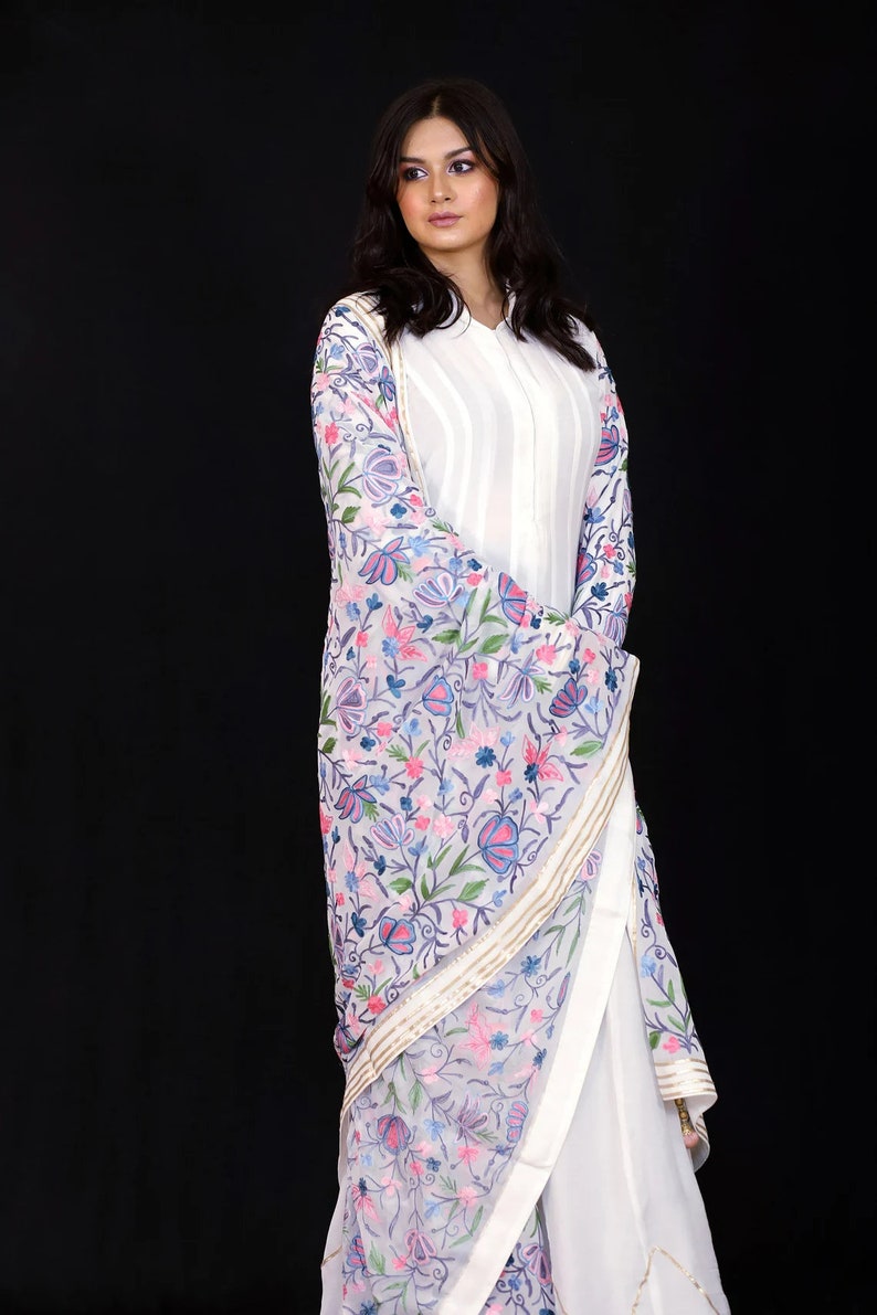 May include: A woman wearing a white long-sleeved shirt with a white dupatta featuring a floral embroidery pattern in shades of pink, blue, and green.