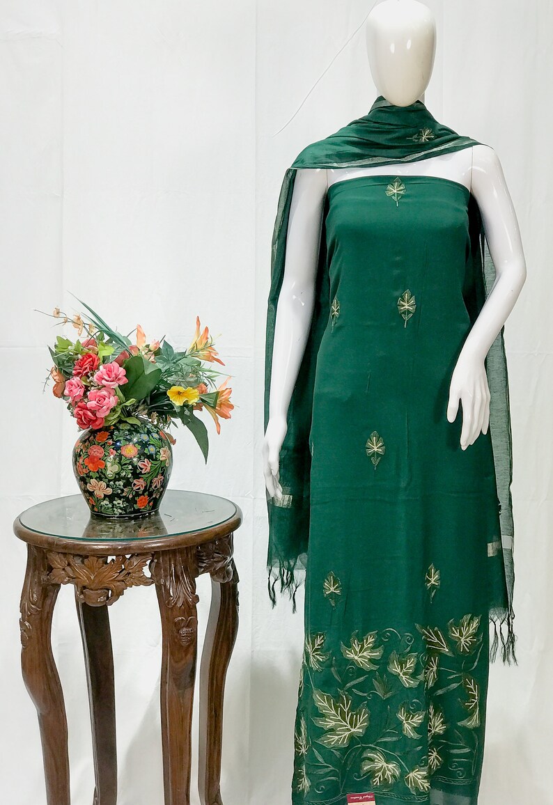 May include: A green cotton dress with gold leaf embroidery. The dress is on a white mannequin and has a matching green dupatta with gold trim.