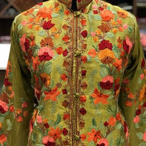 May include: An olive green jacket featuring a high mandarin collar and long sleeves, adorned with intricate floral embroidery in shades of orange, red, and pink. The front closure consists of fabric-covered buttons aligned vertically along the center.