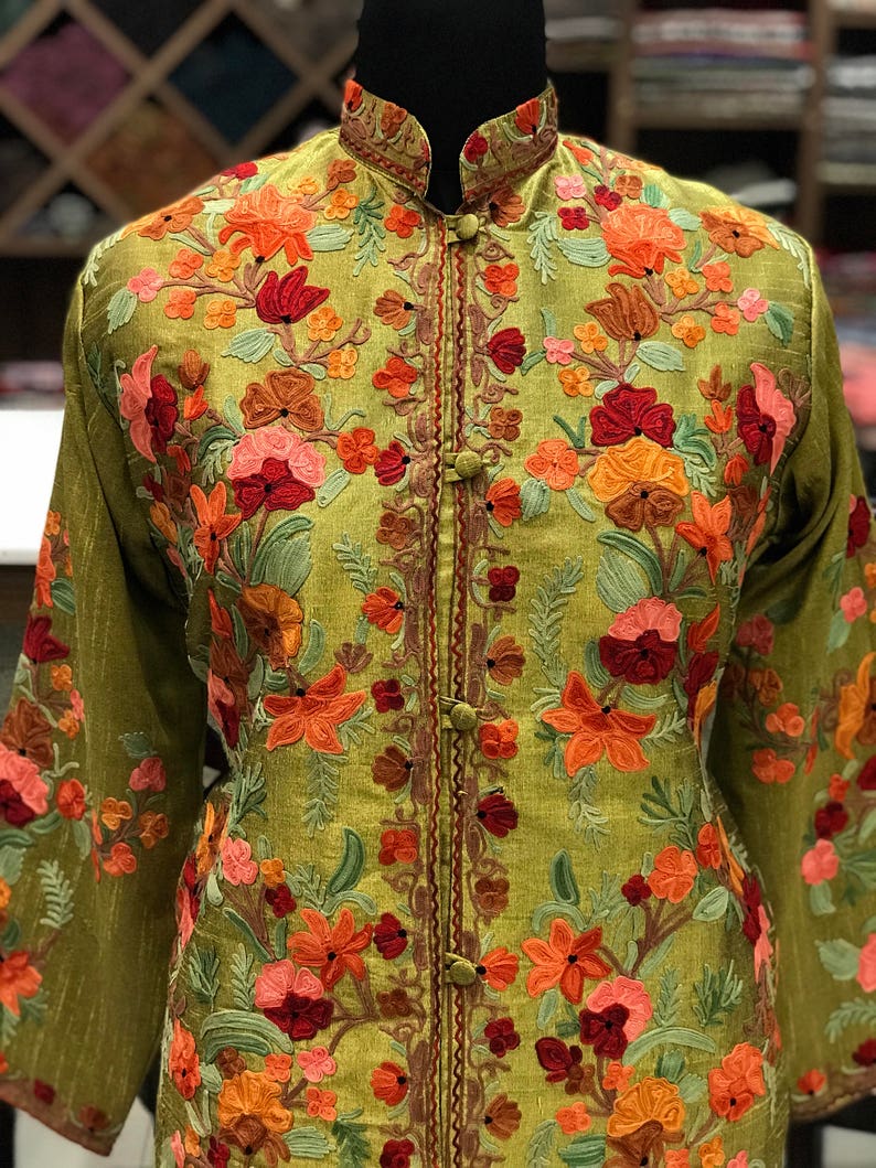 May include: An olive green jacket featuring a high mandarin collar and long sleeves, adorned with intricate floral embroidery in shades of orange, red, and pink. The front closure consists of fabric-covered buttons aligned vertically along the center.