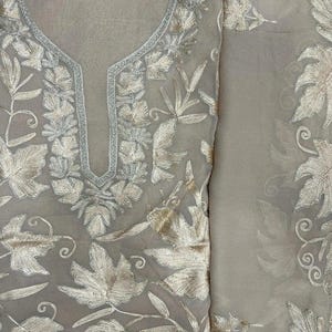 May include: A beige sheer fabric garment featuring intricate metallic gold thread embroidery in a leaf and vine pattern. The neckline is accented with a silver-toned border, and the embroidery covers the front panel and the adjacent fabric section.