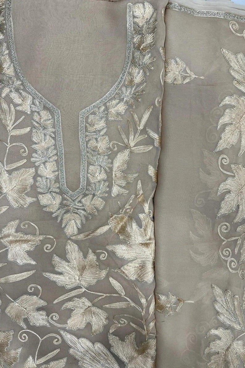 May include: A beige sheer fabric garment featuring intricate metallic gold thread embroidery in a leaf and vine pattern. The neckline is accented with a silver-toned border, and the embroidery covers the front panel and the adjacent fabric section.