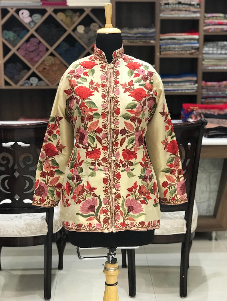 K&ouml;nnte beinhalten: Eine cremefarbene Jacke mit Stehkragen und langen &Auml;rmeln. Die Jacke ist mit einem floralen Muster in Rot-, Rosa- und Gr&uuml;nt&ouml;nen bestickt.