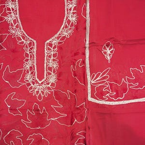 May include: A vibrant red fabric set featuring intricate white floral embroidery. The garment includes a tunic with a U-shaped neckline bordered by detailed stitching and a matching piece of fabric with a single embroidered leaf motif and trim.