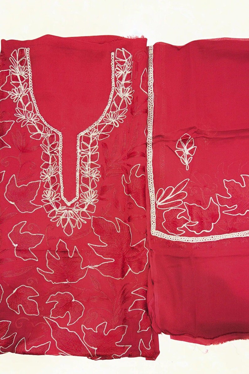 May include: A vibrant red fabric set featuring intricate white floral embroidery. The garment includes a tunic with a U-shaped neckline bordered by detailed stitching and a matching piece of fabric with a single embroidered leaf motif and trim.