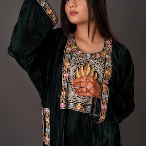 May include: A dark green velvet tunic featuring a round neckline and long sleeves. The garment is adorned with intricate floral embroidery in shades of pink, yellow, and blue along the cuffs, the front panel, and the side slits.