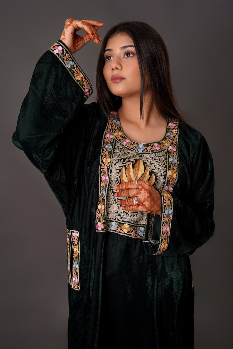 May include: A dark green velvet tunic featuring a round neckline and long sleeves. The garment is adorned with intricate floral embroidery in shades of pink, yellow, and blue along the cuffs, the front panel, and the side slits.