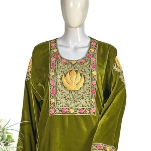 May include: An olive green velvet tunic featuring a scoop neckline and long sleeves. The chest area is adorned with intricate gold, pink, and yellow floral embroidery surrounding a central golden leaf motif. Similar floral embroidery accents the cuffs.