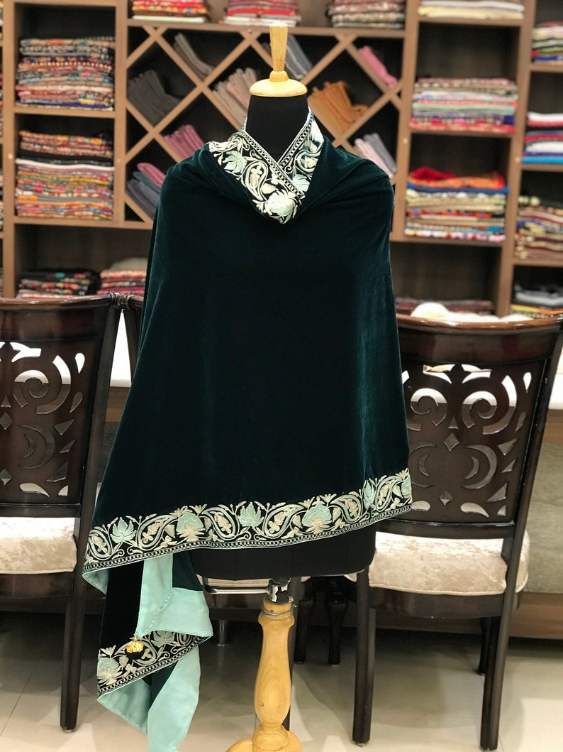 May include: A dark green velvet shawl with a light green and white embroidered border. The shawl is draped over a mannequin and has a light green lining.