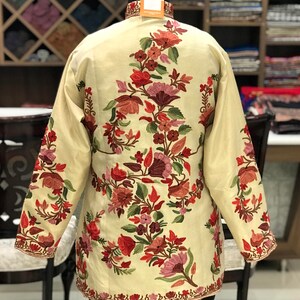 K&ouml;nnte beinhalten: Eine cremefarbene Jacke mit einem floralen Stickmuster. Die Stickerei zeigt rote, rosa und gr&uuml;ne Blumen.