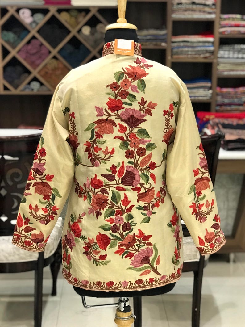K&ouml;nnte beinhalten: Eine cremefarbene Jacke mit einem floralen Stickmuster. Die Stickerei zeigt rote, rosa und gr&uuml;ne Blumen.