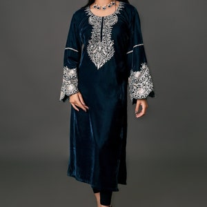 May include: A long, dark teal velvet dress with intricate white embroidery around the neckline and cuffs. The dress has long sleeves and a flowing silhouette. The model is wearing black leggings and black heels.