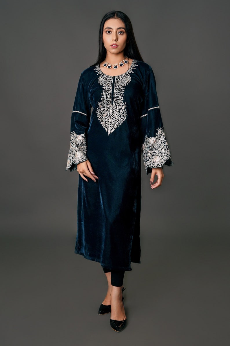 May include: A long, dark teal velvet dress with intricate white embroidery around the neckline and cuffs. The dress has long sleeves and a flowing silhouette. The model is wearing black leggings and black heels.