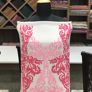 May include: A white fabric panel displayed on a mannequin, featuring intricate embroidery in shades of light pink and dark pink. The embroidery consists of symmetrical, swirling floral and vine-like patterns that extend vertically along the sides.