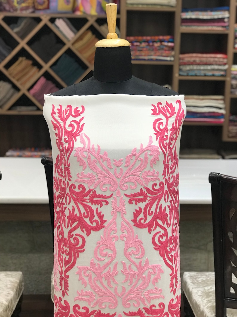 May include: A white fabric panel displayed on a mannequin, featuring intricate embroidery in shades of light pink and dark pink. The embroidery consists of symmetrical, swirling floral and vine-like patterns that extend vertically along the sides.