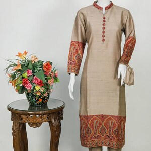 May include: A beige silk kurta suit with intricate embroidery in red, orange, and gold. The long-sleeved tunic features detailed patterns on the cuffs and hem, paired with matching trousers. The suit is displayed on a mannequin.