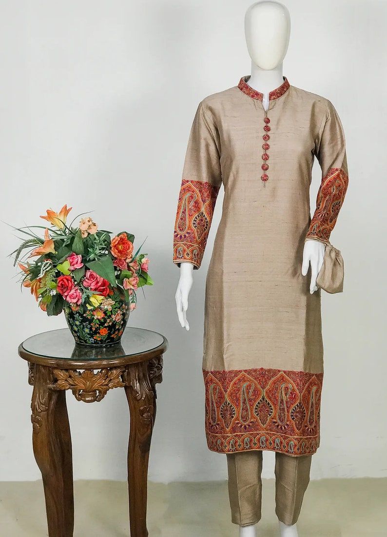 May include: A beige silk kurta suit with intricate embroidery in red, orange, and gold. The long-sleeved tunic features detailed patterns on the cuffs and hem, paired with matching trousers. The suit is displayed on a mannequin.
