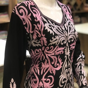 May include: A black long-sleeved tunic featuring a V-neckline and intricate embroidery in shades of light pink and silver. The floral-inspired scrollwork pattern covers the front bodice and extends down the sleeves, creating a detailed contrast.
