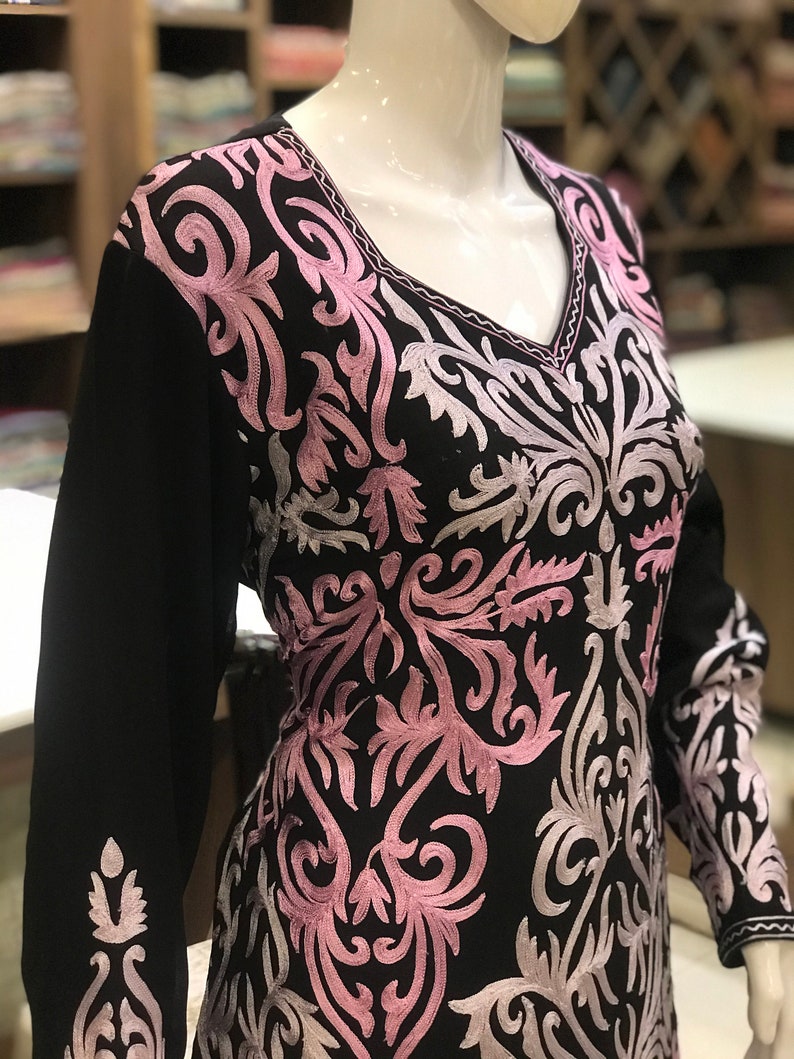 May include: A black long-sleeved tunic featuring a V-neckline and intricate embroidery in shades of light pink and silver. The floral-inspired scrollwork pattern covers the front bodice and extends down the sleeves, creating a detailed contrast.