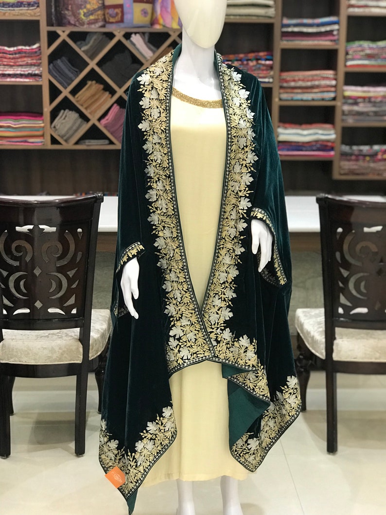 May include: A dark green velvet shawl with intricate white and gold embroidery. The shawl is draped over a mannequin wearing a cream-colored dress.