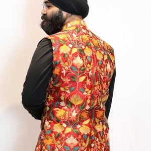 Op de afbeelding: Een rood vest met een bloemenpatroon in tinten geel, groen en blauw. Het vest is geborduurd met ingewikkelde details en heeft een mandarijnskraag.