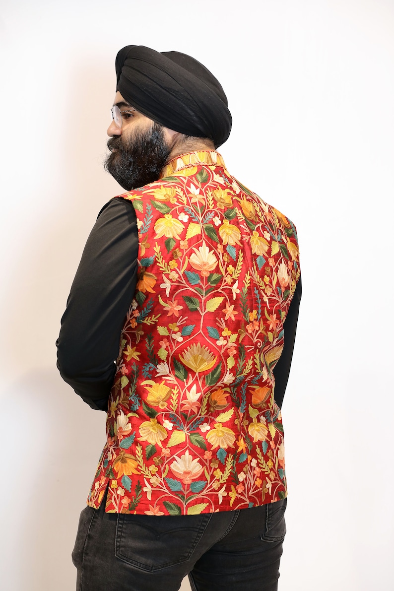Op de afbeelding: Een rood vest met een bloemenpatroon in tinten geel, groen en blauw. Het vest is geborduurd met ingewikkelde details en heeft een mandarijnskraag.