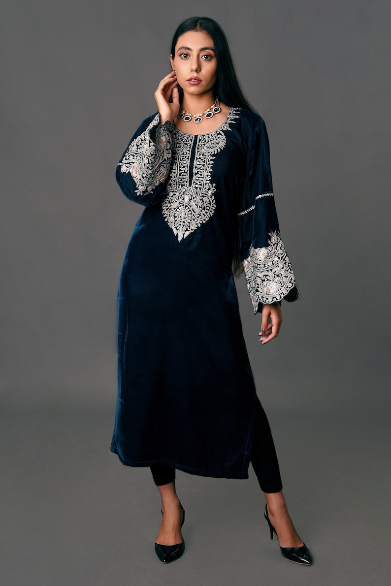 May include: A long, dark blue velvet tunic with intricate silver embroidery around the neckline and on the flared sleeves. The tunic is paired with black leggings and heels. The overall style is elegant and sophisticated.