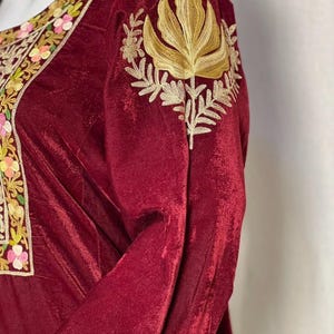 May include: A deep burgundy velvet long-sleeve garment featuring intricate gold thread embroidery on the shoulder and a neckline border decorated with colorful floral patterns in shades of pink, yellow, and green on a cream-colored fabric base.