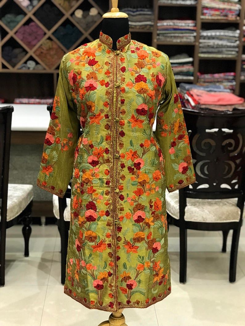 May include: A long-sleeved olive green tunic featuring a mandarin collar and a front button closure. The garment is decorated with intricate floral embroidery in shades of orange, red, pink, and brown, covering the front and sleeves.