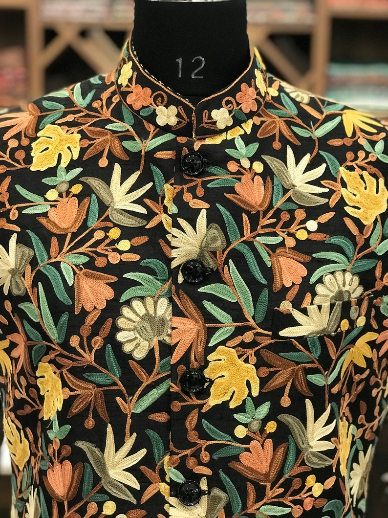 K&ouml;nnte beinhalten: Eine schwarze Weste mit einem floralen Stickmuster in Gr&uuml;n-, Braun- und Gelbt&ouml;nen. Die Weste hat einen Stehkragen und f&uuml;nf Kn&ouml;pfe.