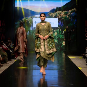 May include: An olive green salwar kameez featuring a long-sleeved tunic with subtle embroidery, matching straight-leg trousers, and a draped shawl adorned with intricate floral patterns in shades of green, gold, and blue, worn with simple sandals.