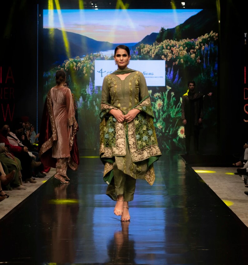 May include: An olive green salwar kameez featuring a long-sleeved tunic with subtle embroidery, matching straight-leg trousers, and a draped shawl adorned with intricate floral patterns in shades of green, gold, and blue, worn with simple sandals.