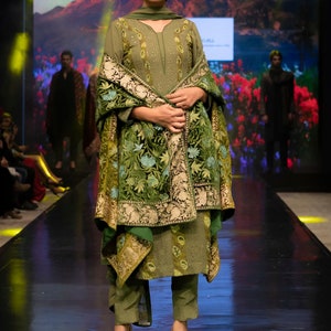 May include: An olive green tunic and trousers set featuring intricate embroidery on the bodice and a matching shawl draped over the shoulders. The shawl displays a dense, multi-coloured floral pattern with gold accents and a wide, ornate border.