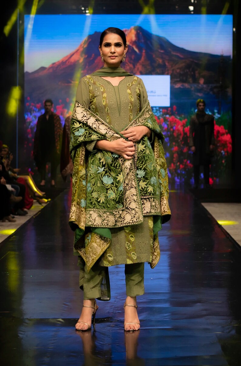 May include: An olive green tunic and trousers set featuring intricate embroidery on the bodice and a matching shawl draped over the shoulders. The shawl displays a dense, multi-coloured floral pattern with gold accents and a wide, ornate border.